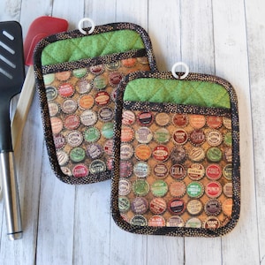 May include: Two green and black pot holders with a pattern of vintage bottle caps. The bottle caps have various colors and designs, including red, blue, yellow, and green.