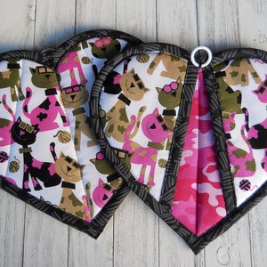 May include: Three heart-shaped pot holders with a black and white cat design. The pot holders have a pink and camouflage pattern on the back.