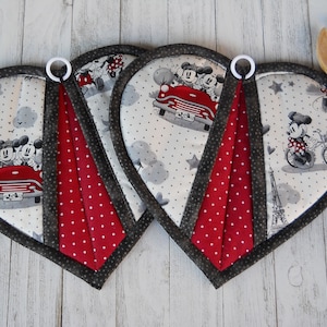 May include: Two heart-shaped pot holders with a red and white polka dot design. The pot holders feature a black and white print of Mickey and Minnie Mouse in a red car. One pot holder has a black and white print of Minnie Mouse riding a bicycle.