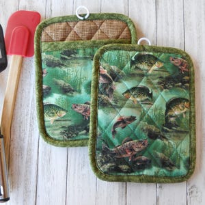 May include: Two green and brown quilted pot holders with a fish print. The pot holders have a loop for hanging.