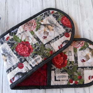 May include: Quilted oven mitts with a floral and dog print. The mitts feature a black and white background with red roses and dog illustrations. The edges are trimmed with a black polka dot fabric. The interior is a deep red.