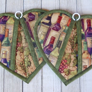 May include: Two heart-shaped pot holders with a wine bottle and grape design. The fabric features various wine bottles in shades of red, green, and brown. The edges are trimmed with a green fabric and have white hanging loops.