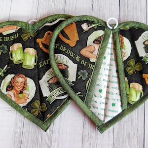 May include: Set of three green and black heart-shaped pot holders with Irish-themed designs. The designs include a pub, a woman drinking beer, a pot of gold, and a harp. The text on the pot holders reads "Eat Drink & Be Irish" and "Authentic Irish Pub".