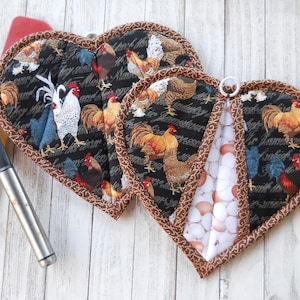 May include: Two heart-shaped fabric potholders featuring a black background with a rooster and hen print, accented by a brown patterned border. One potholder includes a white and brown egg pattern insert. A black slotted spatula is positioned nearby.