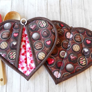 May include: Two heart-shaped pot holders with a chocolate and pink pattern. The pot holders are made of fabric and have a loop for hanging.