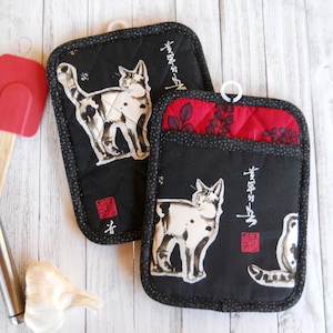 May include: Two black pot holders with a white cat illustration and Japanese calligraphy. One pot holder has a red quilted pocket. Kitchen utensils and garlic are in the background.