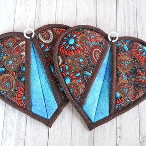 May include: Two heart-shaped quilted potholders. The potholders feature a paisley pattern in red, turquoise, and brown. A section of each potholder is a solid turquoise color. Each potholder has a brown border and a white hanging loop.