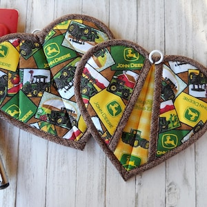 May include: Two brown heart-shaped pot holders with a brown trim. The pot holders are made of fabric with a patchwork design featuring John Deere tractors and logos in green, yellow, and brown.