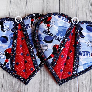 May include: Three heart-shaped pot holders with a hockey theme. The pot holders are red and blue with a white star pattern. The front of the pot holders features a blue and white hockey player pattern with the words "Power Play" and "Central".
