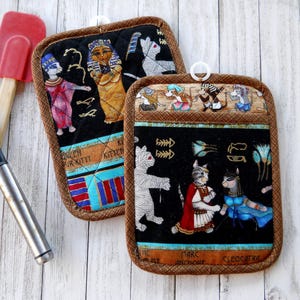May include: Two quilted fabric pot holders featuring Egyptian-themed cat illustrations on a black background with brown trim. One pot holder displays cats dressed as royalty, while the other shows cats in ancient attire with text reading Marc Anchoue and Cleocatra.