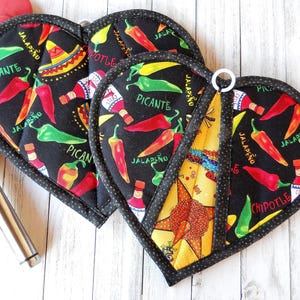 May include: Heart-shaped black pot holders with a colorful chili pepper and sombrero print. The pot holders have a white hanging loop and are trimmed with a black and white polka dot border. The words "PICANTE", "JALAPENO", and "CHIPOTLE" are visible.