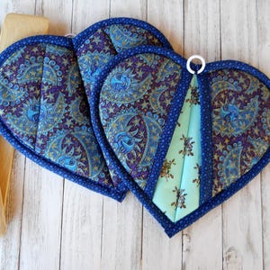 May include: Heart-shaped quilted pot holders with a blue paisley pattern and a light blue floral accent. The pot holders have a dark blue border and a white hanging loop. Wooden cooking utensils are visible in the background.
