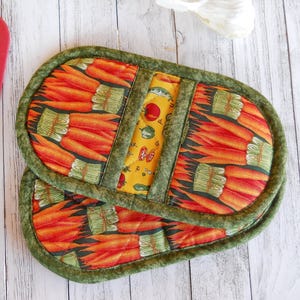 May include: Two quilted, oval-shaped pot holders. The main pattern features orange carrots with green tops on a black background. A yellow strip with vegetable illustrations divides the two pot holders. The edges are trimmed with green fabric.