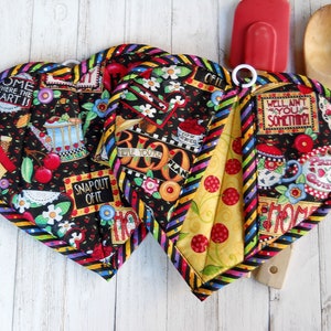 May include: Two heart-shaped fabric pot holders featuring a black background with colorful illustrations of cherries, houses, and text including "HOME WHERE THE HEART IS," "SNAP OUT OF IT," and "WELL AIN'T YOU SOMETHIN'!" with striped black and yellow trim.