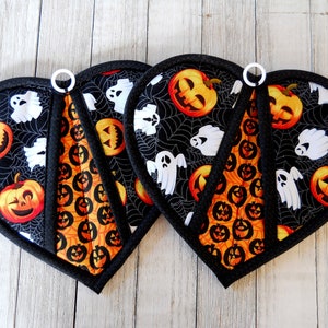 May include: Two black heart-shaped pot holders with a Halloween theme. The pot holders feature a black background with orange pumpkins, white ghosts, and spiderwebs. The pot holders have a white loop handle.
