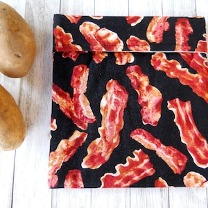 May include: A black fabric reusable snack bag with a red and white bacon print. Two brown potatoes are in the background.