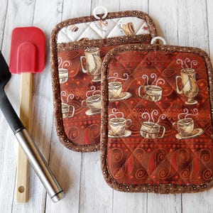 May include: Two brown and white quilted pot holders with a coffee cup pattern. The pot holders have a brown trim and a white background with brown coffee cups and steam.