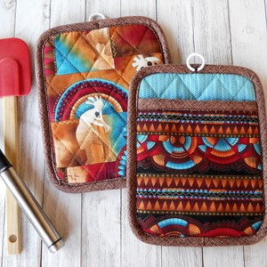 May include: Two rectangular quilted pot holders with brown fabric trim and white hanging loops. One features a Kokopelli figure in orange and blue tones, while the other displays a geometric pattern in blue, red, and black. A red spatula is included.