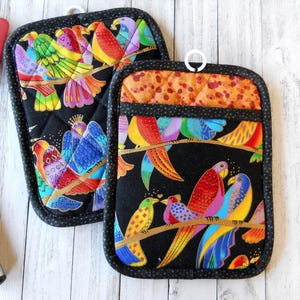 May include: Two quilted pot holders with a black background and a colorful bird design. The birds are perched on a branch, with vibrant feathers in shades of red, yellow, blue, and green. Each pot holder has a hanging loop.