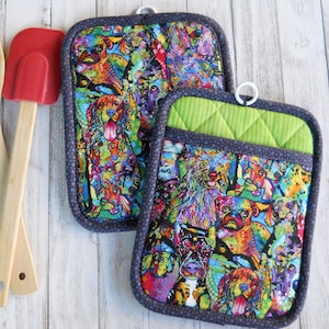 May include: Two rectangular pot holders with a colorful dog print, featuring a variety of breeds in vibrant hues. The pot holders have a gray border with a polka dot pattern and a green accent. A wooden spoon and red spatula are also in the image.