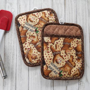 May include: Two rectangular quilted pot holders featuring a repeating pattern of gingerbread people, red berries, and the text GINGERBREAD, with brown plaid trim and a white hanging loop, alongside a black slotted turner and a red spatula.