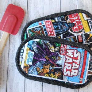 May include: Two oval-shaped Star Wars themed pot holders. The fabric features comic book panels with Darth Vader and other characters. The edges are trimmed with a black and white polka dot fabric. A red spatula and black spatula are in the background.