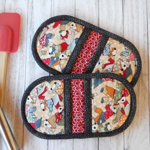 May include: Two oval-shaped quilted pot holders. The main fabric features a pattern of dogs in various outfits on a beige background. A red fabric with white polka dots and a black fabric with white polka dots are used as accents.