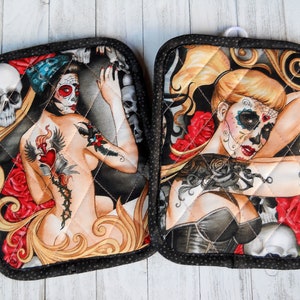 May include: Two quilted oven mitts featuring Day of the Dead themed artwork. Each mitt displays a woman with sugar skull makeup, surrounded by roses and skulls. The mitts have a black border with white polka dots.