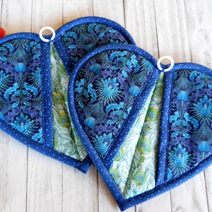 May include: Two heart-shaped quilted pot holders. The pot holders feature a blue floral pattern with a blue border. Each pot holder has a white loop for hanging. A red spatula is visible on the left.