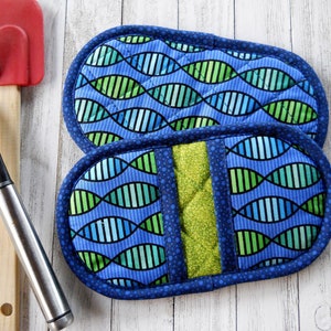 May include: Two blue and green quilted oven mitts with a DNA-like pattern. The mitts have a dark blue border with small blue dots. A red spatula and a black spatula are visible in the background.