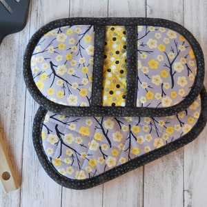 May include: Two quilted, oval-shaped pot holders. The main fabric is gray with a yellow floral pattern and black branches. A yellow and black polka dot panel is centered between the two pot holders. The edges are trimmed with a dark gray dotted fabric.