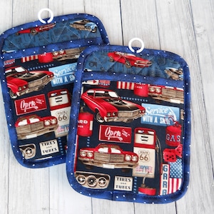 May include: A pair of blue and white pot holders with a red, white, and blue car and gas station theme. The pot holders have a loop for hanging. The text on the pot holders reads "Open", "Service with a Smile", "Route 66", "Tires and Tubes", and "Gas".