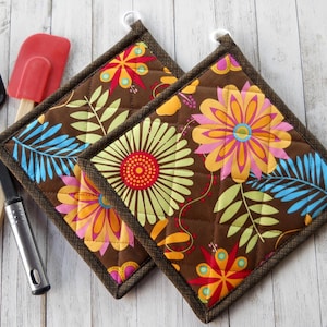 May include: Two square quilted pot holders featuring a brown background with a vibrant floral and leaf pattern in shades of pink, orange, yellow, lime green, and blue. Each pot holder has a dark brown fabric border and a small white hanging loop.