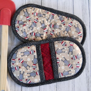 May include: Two quilted, oval-shaped pot holders. The main fabric features a repeating pattern of sock monkey figures on a beige background with white buttons. A red stripe with a small dot pattern is centered on one. The edges are trimmed with a dark gray fabric.