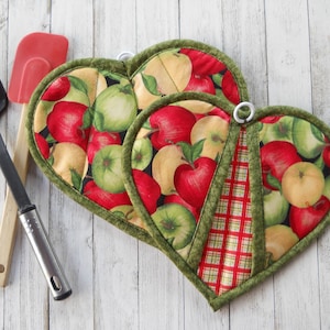 May include: Two heart-shaped fabric pot holders featuring a pattern of red, green, and yellow apples on a black background, with one featuring a red and green plaid section. A black slotted turner and a red silicone spatula are positioned nearby.