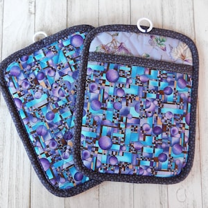 May include: Two blue and purple quilted pot holders with a geometric pattern. The pot holders have a black trim and a white loop for hanging.
