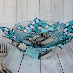 May include: A clear glass bowl nested in a teal and white polka dot and black and white patterned bowl cozy. The cozy has a light blue interior with a vintage illustration. A fork and spoon are in the foreground.