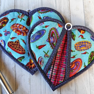 May include: Two heart-shaped blue pot holders with a colorful Mexican-inspired design featuring sombreros and chili peppers. The pot holders have a dark blue trim.