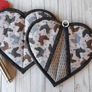 May include: Two heart-shaped pot holders with a rooster print. The pot holders have a black trim and a gray and brown honeycomb pattern on the back.