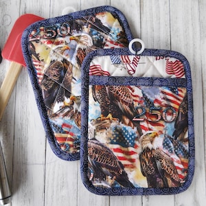 May include: Two patriotic quilted pot holders with a blue border. The fabric features American flags, eagles, and the number 250. One pot holder has a pocket. A black spatula and red spatula are in the background.