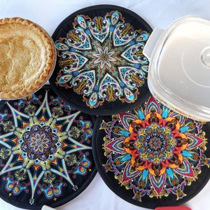 May include: A baked pie with a golden crust sits next to three black, round pot holders with colorful, intricate mandala designs. A clear, square glass dish with a white rim is also present.