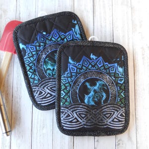 May include: Two black quilted pot holders with a blue and teal dragon design. The pot holders have a diamond pattern and a decorative border. A red spatula and a black spatula are in the background.