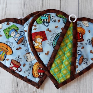 May include: Three heart-shaped pot holders with a brown trim. The pot holders are made of fabric with a blue background and a coffee cup design. One pot holder has the word "Cappuccino" written on it.