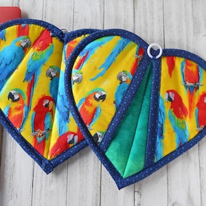 May include: Two heart-shaped oven mitts with a parrot print. The mitts are yellow with blue and red parrots. The mitts have a blue trim and a white loop for hanging.
