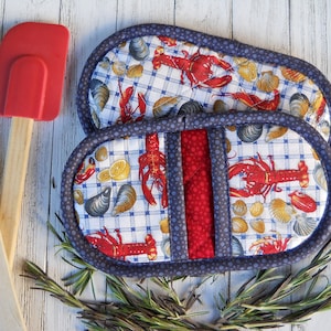 May include: A pair of blue and red oven mitts with a lobster and seashell pattern. The mitts are made of fabric and have a quilted design.