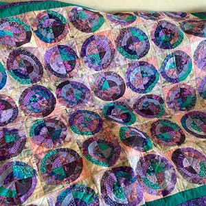 May include: A quilt featuring a repeating pattern of circular motifs in shades of purple, teal, and green. Each circle is composed of smaller fabric segments, set against a light cream background with a solid dark green border trim.