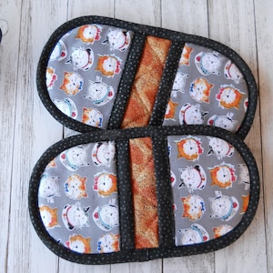 May include: A pair of oval-shaped oven mitts featuring a gray fabric with a repeating pattern of cartoon cat faces in orange, white, and black. Each mitt has a central vertical strip of metallic orange fabric and black polka-dot trim.