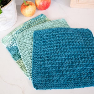 May include: Three square knitted washcloths arranged in a stack, featuring a textured stitch pattern. The cloths display a gradient of colors, ranging from a deep teal to a lighter mint green and a soft sage green, creating a cohesive color palette.