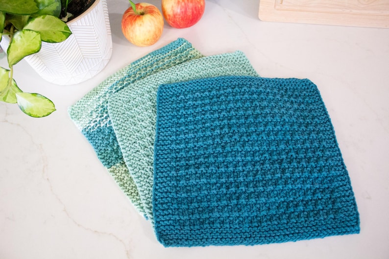 May include: Three square knitted washcloths arranged in a stack, featuring a textured stitch pattern. The cloths display a gradient of colors, ranging from a deep teal to a lighter mint green and a soft sage green, creating a cohesive color palette.