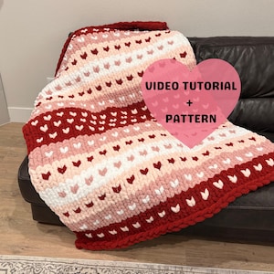 Può includere: Una coperta a maglia grossa nei toni del rosso, rosa e bianco, decorata con motivi a cuore. La coperta è drappeggiata su un divano in pelle marrone scuro. L'immagine include il testo "VIDEO TUTORIAL + PATTERN".